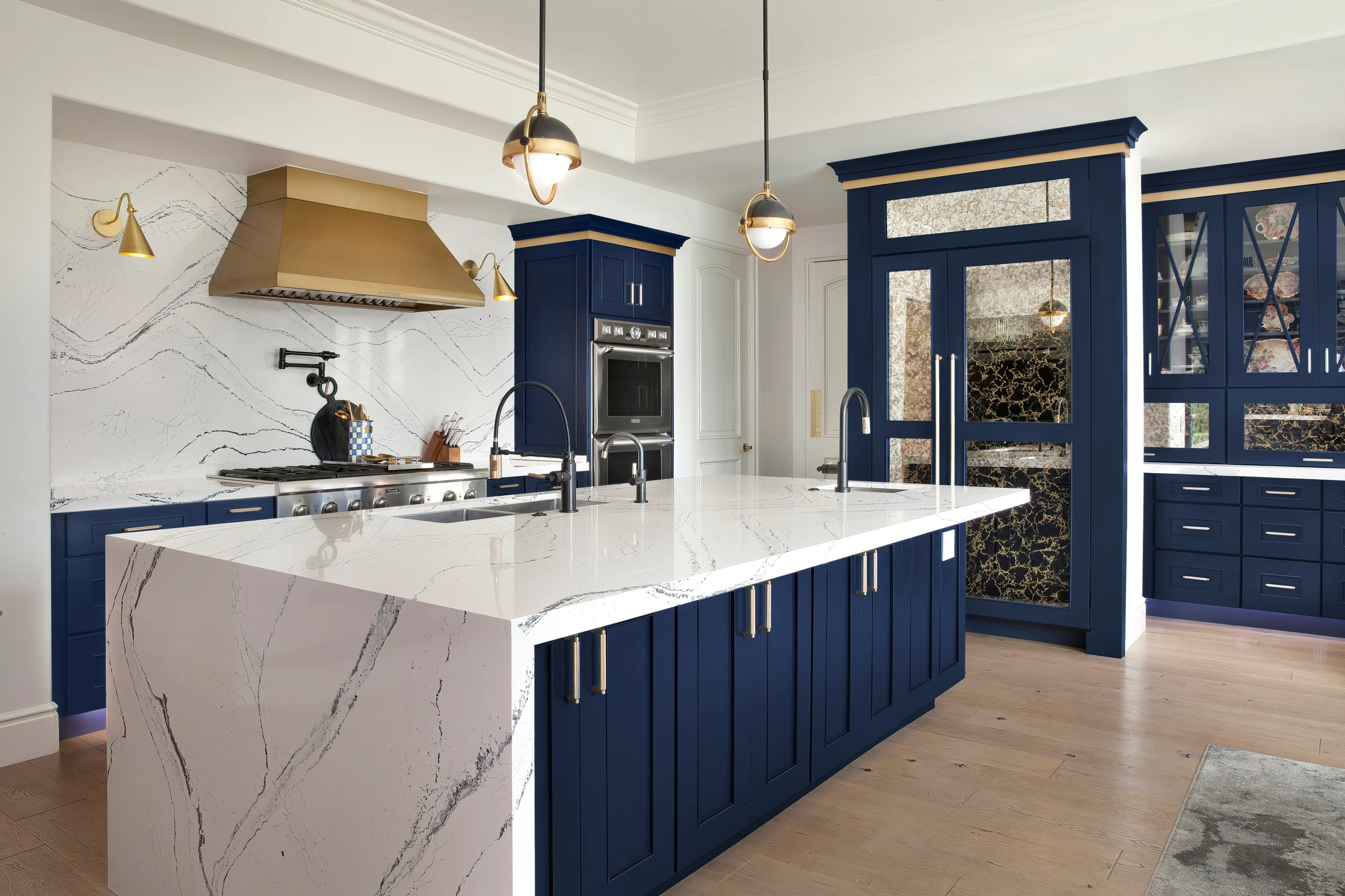 Blue kitchen with white veined island, brass hood, and glass-front cabinetry