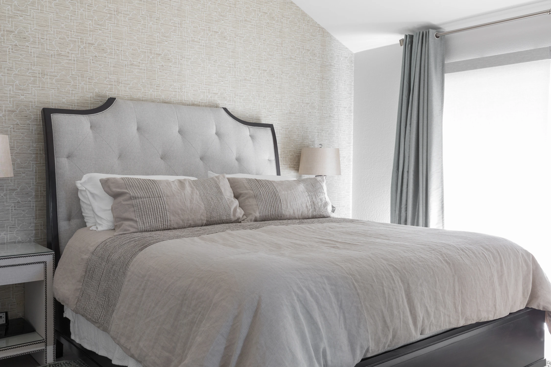 Bedroom bed detail with pale gray headboard, layered bedding, and dark drapery