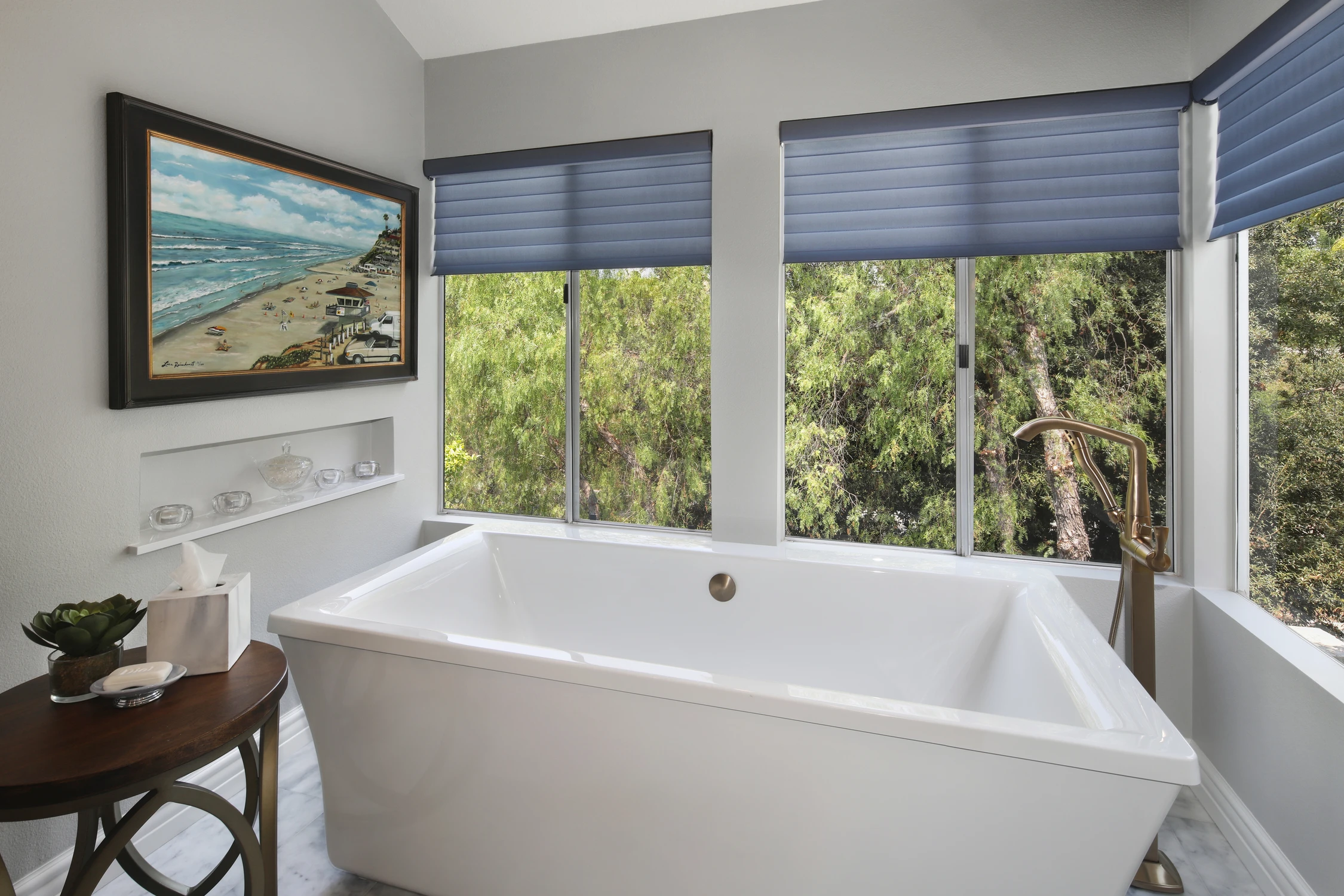 Freestanding bathroom tub beside wide windows and a coastal landscape painting