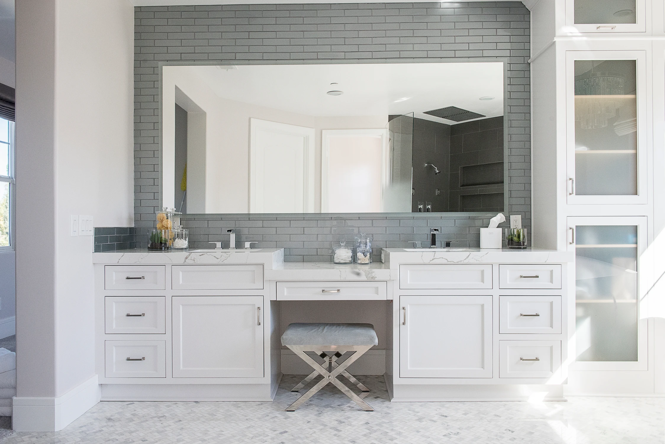 Primary bathroom vanity with white cabinetry, gray tile, and upholstered bench