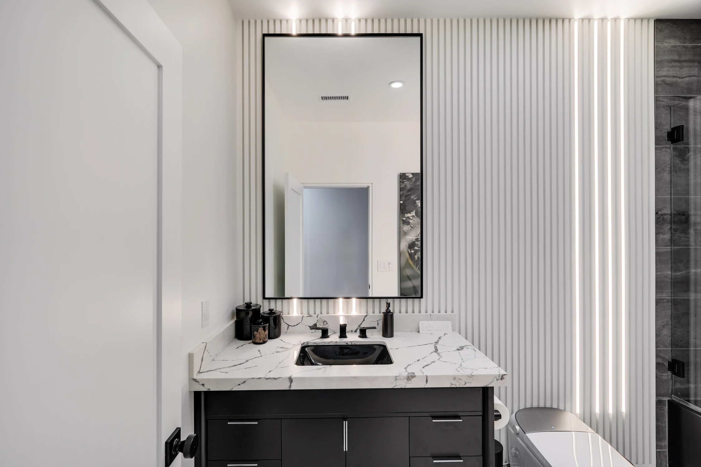 Black bathroom vanity with fluted wall, marble counter, and illuminated mirror