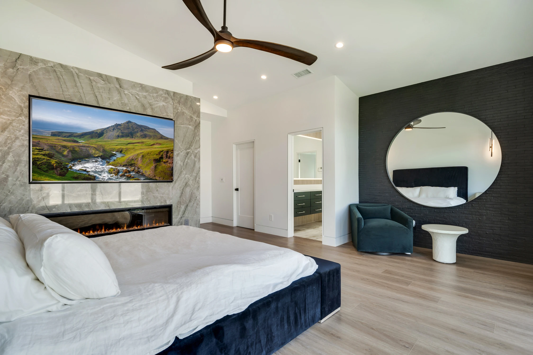 Primary bedroom with black brick accent wall, blue chair, and round mirror