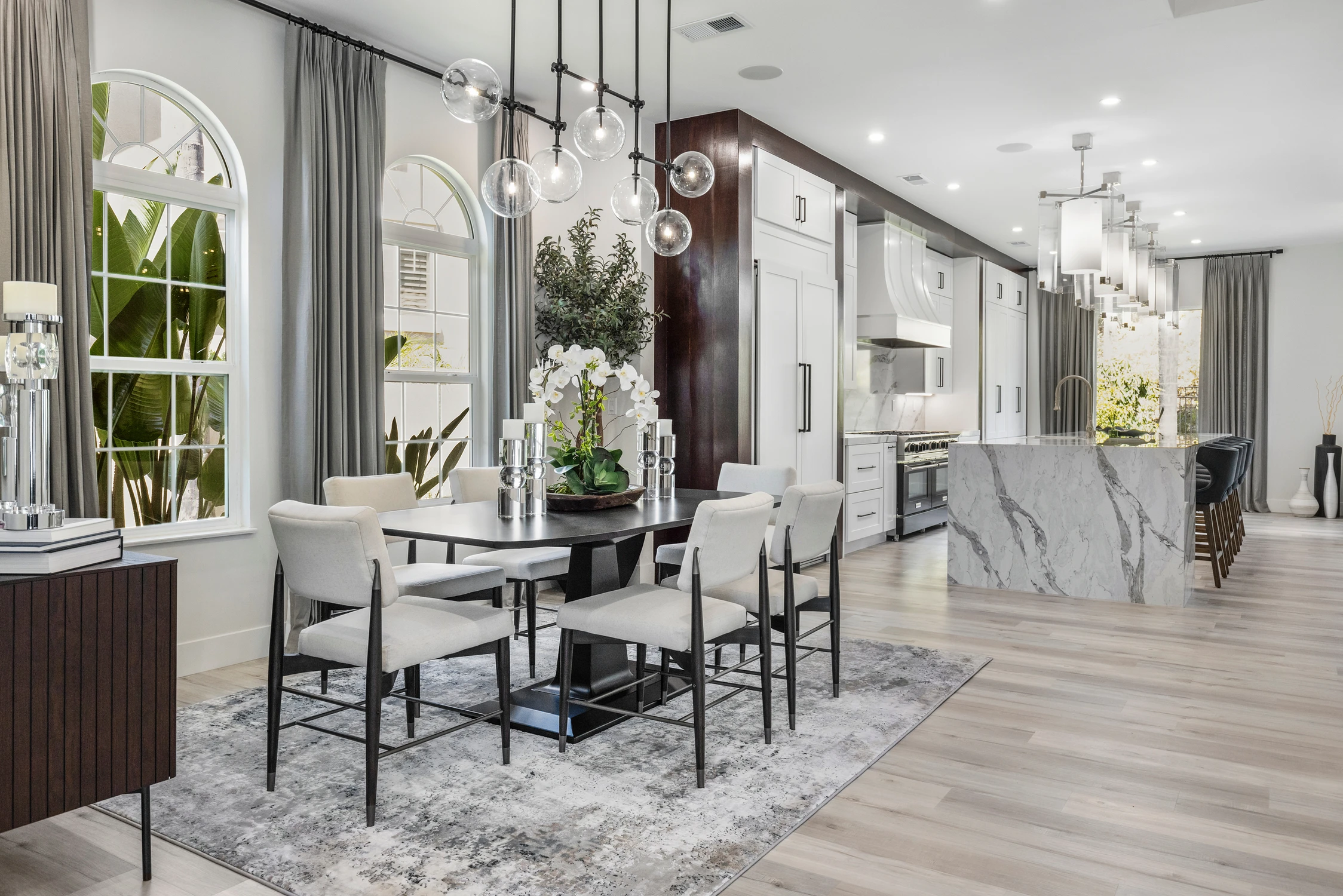 Dining table beneath dark chandelier with view toward bright kitchen